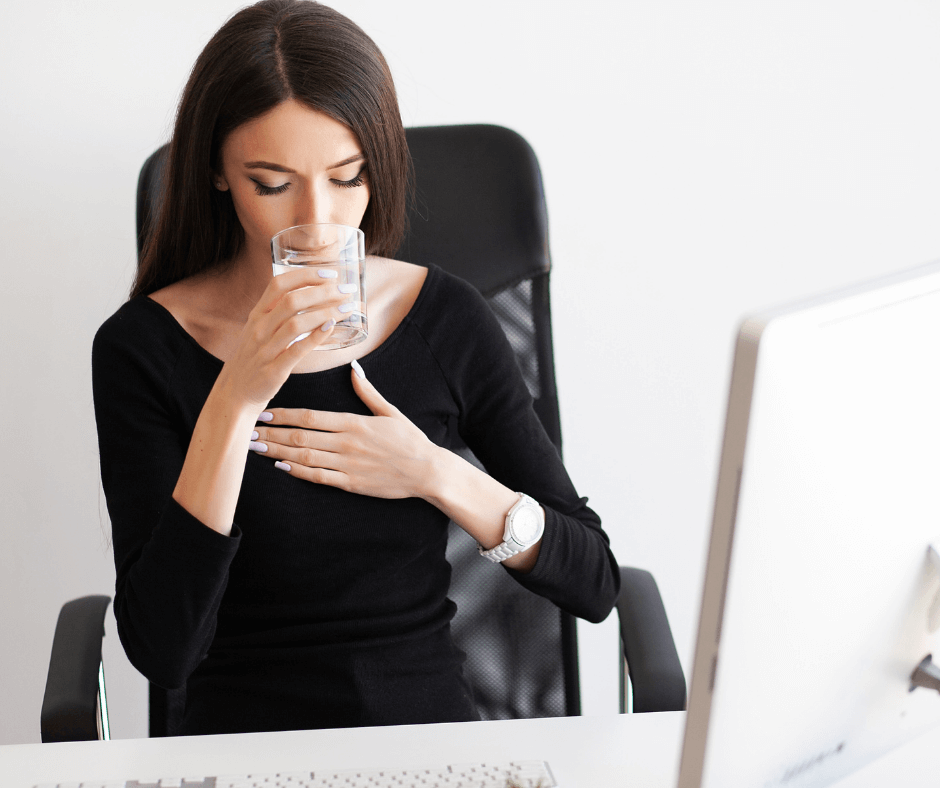 A white woman in a black shirt sits at her desk drinking a glass of water with her other hand on her chest