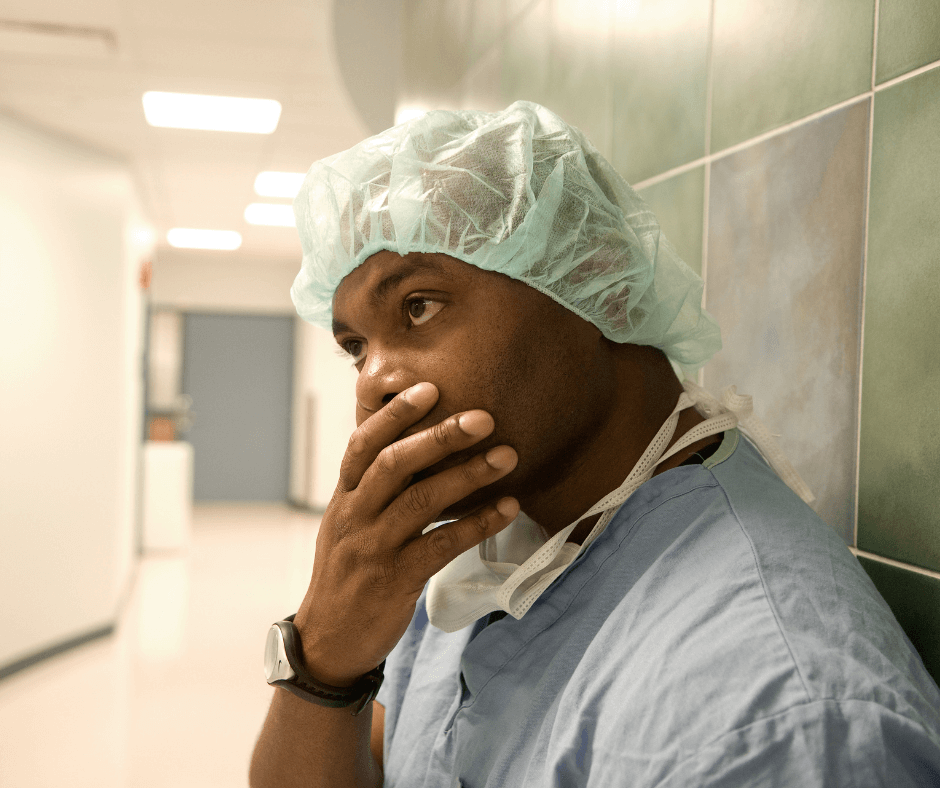 A black male doctor stands in scrubs with a scrub cap, leaning against a wall with his hand over his mouth looking distressed