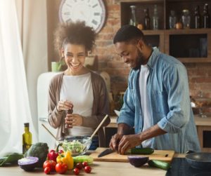 a black male and a black female stand at a kitchen island smiling while they cut vegetables for a salad, the bowl is in front of them