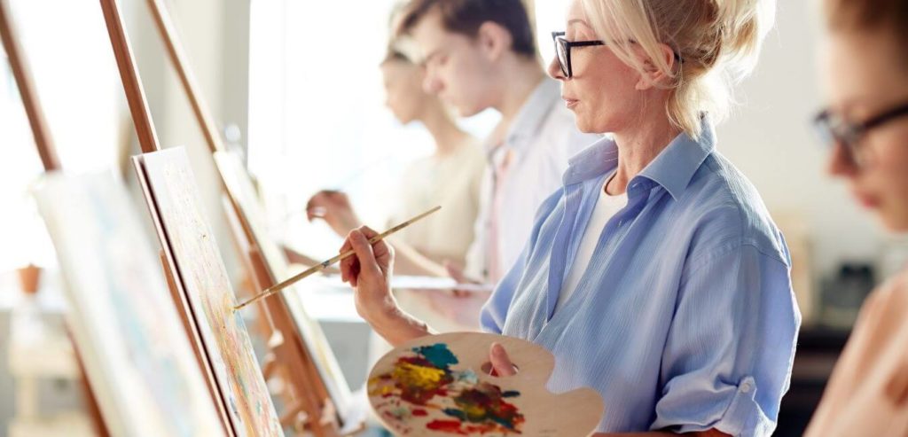 A white woman in a blue shirt paints at an easel. She is in a line of other artists on either side.