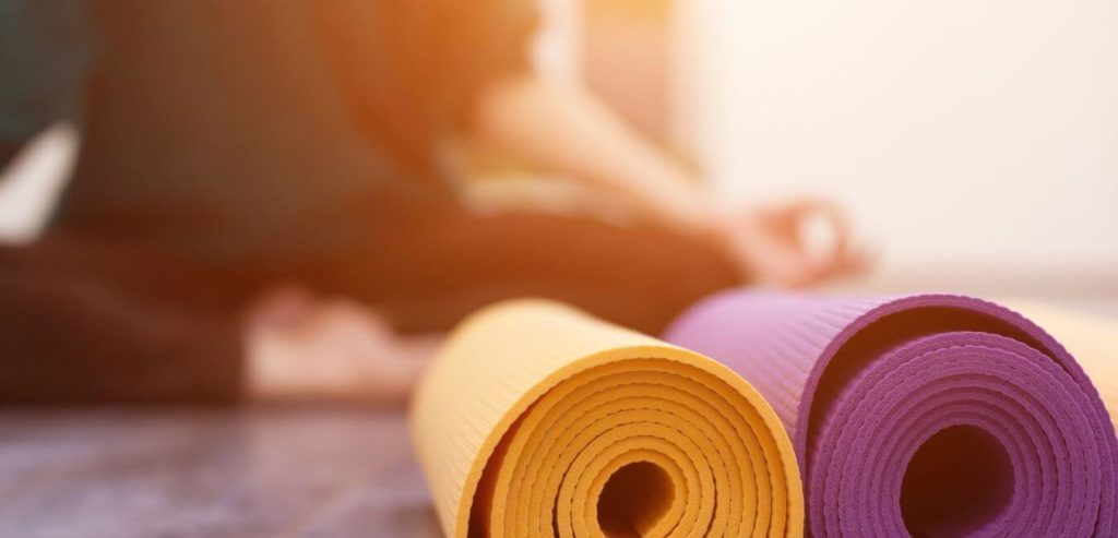 two yoga mats rolled up in the foreground, one yellow and one magenta. A female is in the blurry background crossed legged and practicing mindfulness 