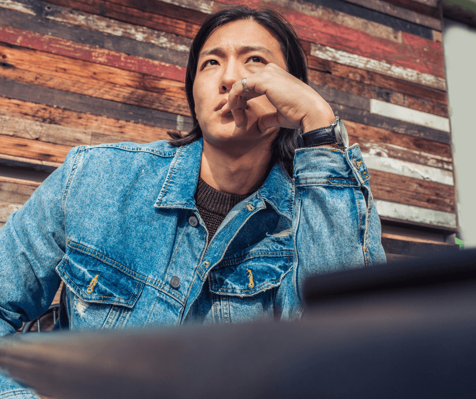 An asian man with shoulder length hair and a jean jacket sits thinking
