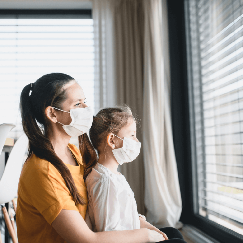 A mother wearing a yellow shirt and wearing a mask hold her elementary aged daughter also wearing a mask. They are both looking out of a window in their home