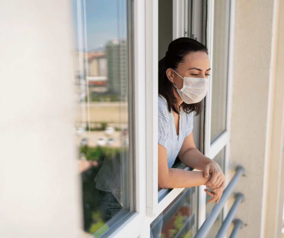 A white woman wearing a mask looks out of a building window