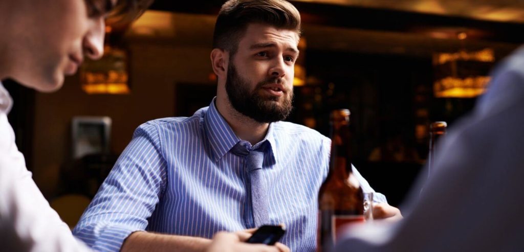 A man in a blue shirt and tie, white with a beard holds a beer and is in discussion