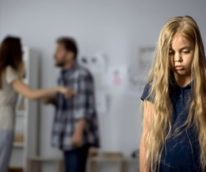 A small girl with long blonde hair is at the front of the camera looking down at the ground with a sad face. In the background, fuzzy couple arguing