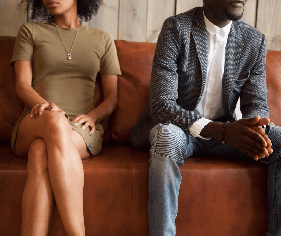 a black woman and black man sit on a rust colored couch. Their heads are not visible but their bodies are turned away from each other