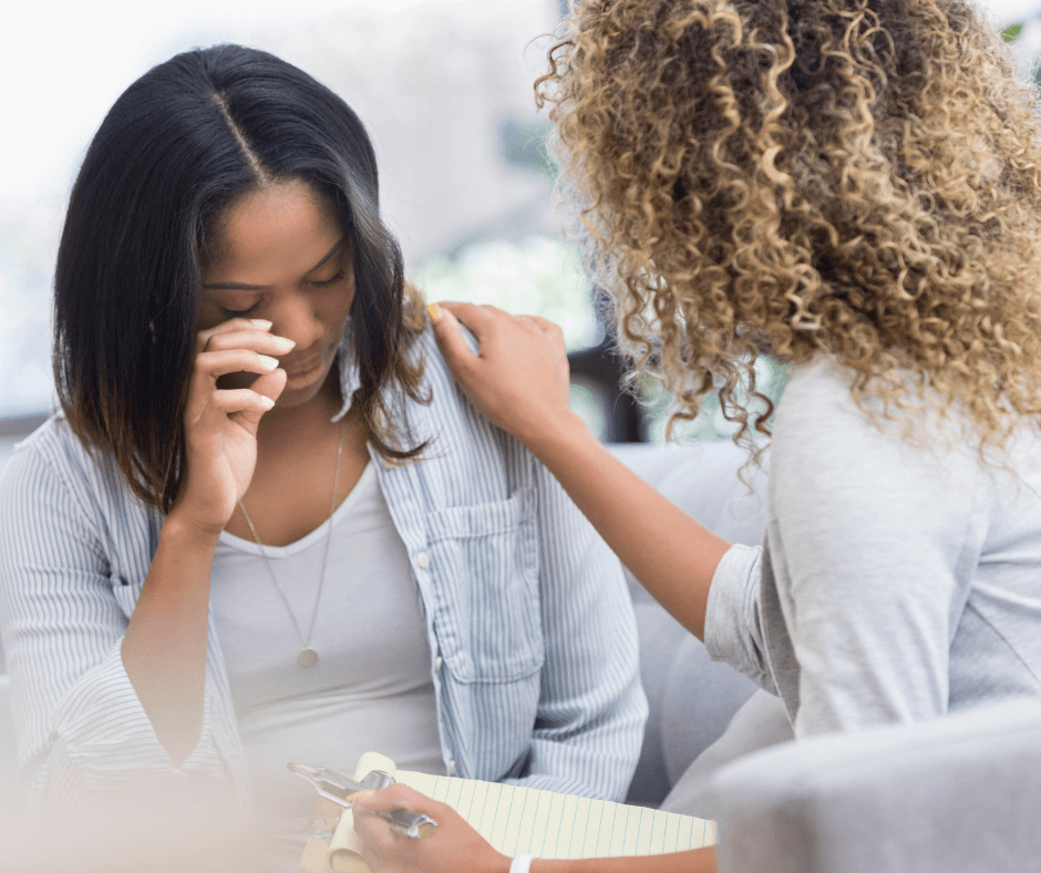 Two women of color sit next to each other, one is crying while the other puts a hand on her shoulder