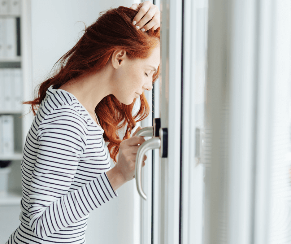 A white woman with red hair and striped shirt stands at a glass door with her hand on the handle looking afraid of going outside