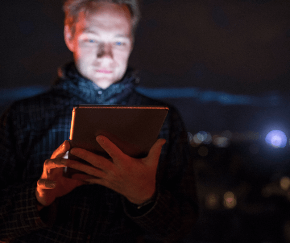 A white man stands in the dark holding his ipad