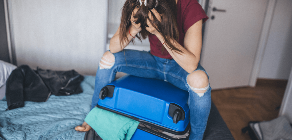 A depressed young woman sitting on suitcase with head in her hands, struggling to close it.