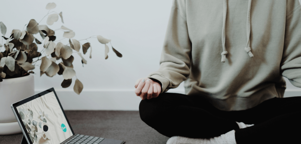 A woman sitting in lotus position besides her laptop and plants while meditating indoors