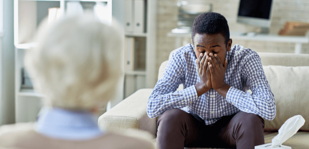 A frustrated black male with both hands on his face sitting on a couch trying to figure problem out to a psychiatrist who is sitting in front of him 