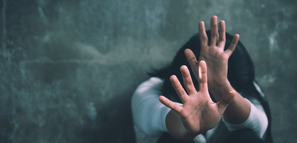 A scared young woman sitting on a floor waving her hands towards an abusive act.