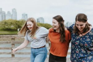 three women walking on brown wooden dock near high rise building during daytime, participating in the levels of intimacy